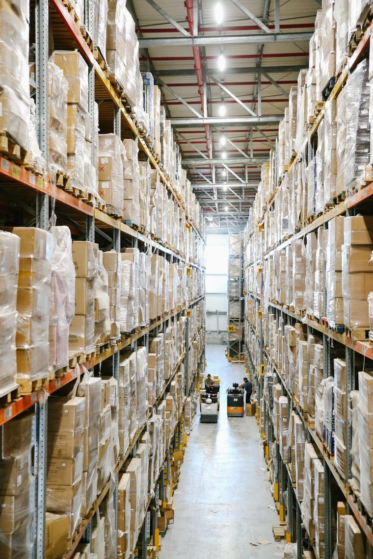Allée d'entrepôt logistique bordée de palettes filmées et de cartons empilés sur plusieurs niveaux.
